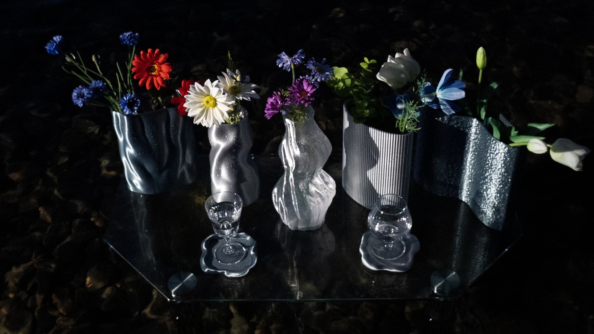 Collection Chrome Core complète avec cinq vases et deux dessous de verre Fuso sur une table en verre, photographiée de nuit dans l’eau vu du dessus