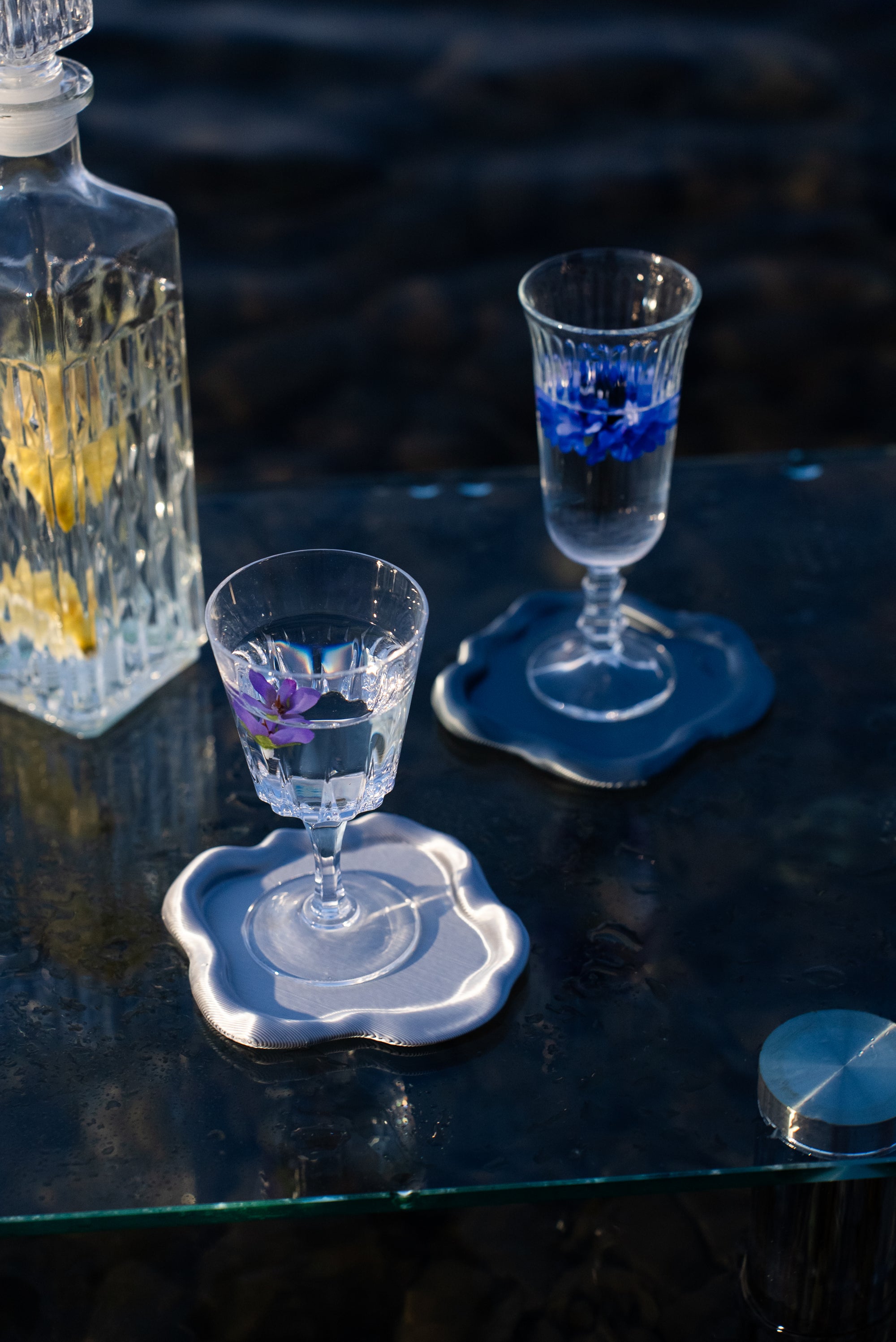 Deux verres posés sur les dessous de verre FUSO de S7DE Studio en finition chrome et aether, sur une table en verre au lever du soleil.