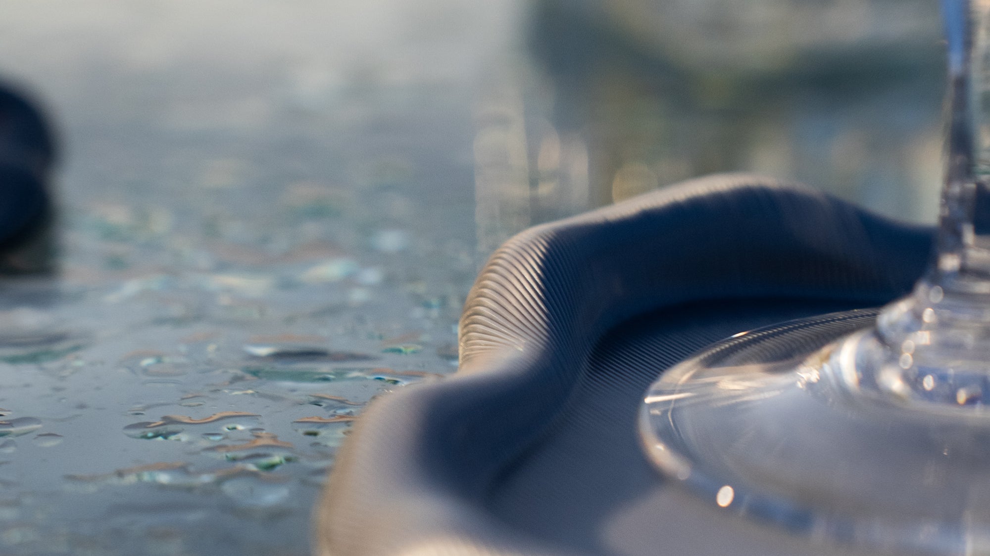 Dessous de verre Fuso en PLA chromé, détail texturé avec verre à pied posé dessus, sur une table mouillée dans la mer