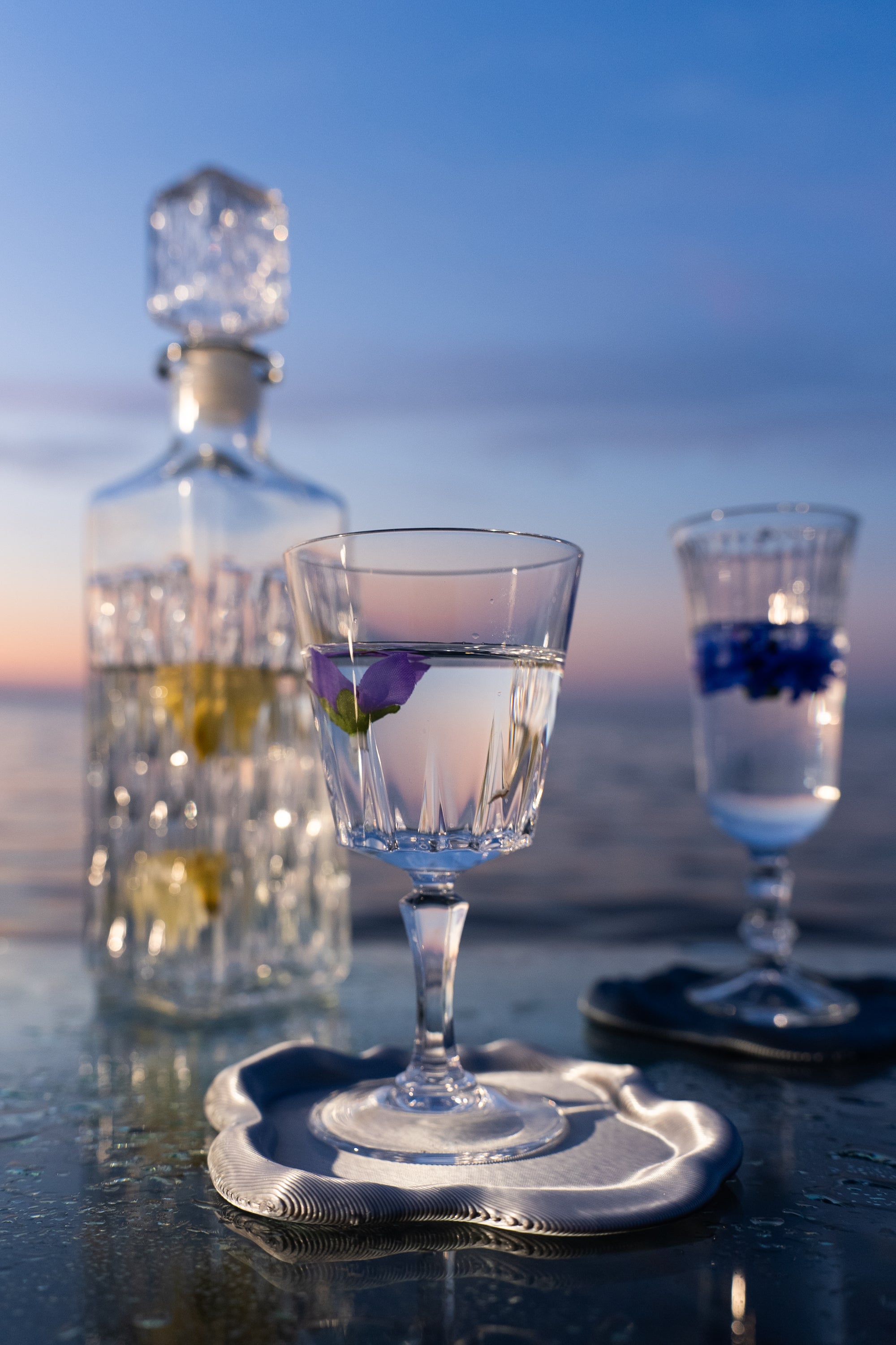 Dessous de verre FUSO de S7DE Studio en finition chrome, utilisé pour un verre à pied, sur une table en verre au lever du soleil.
