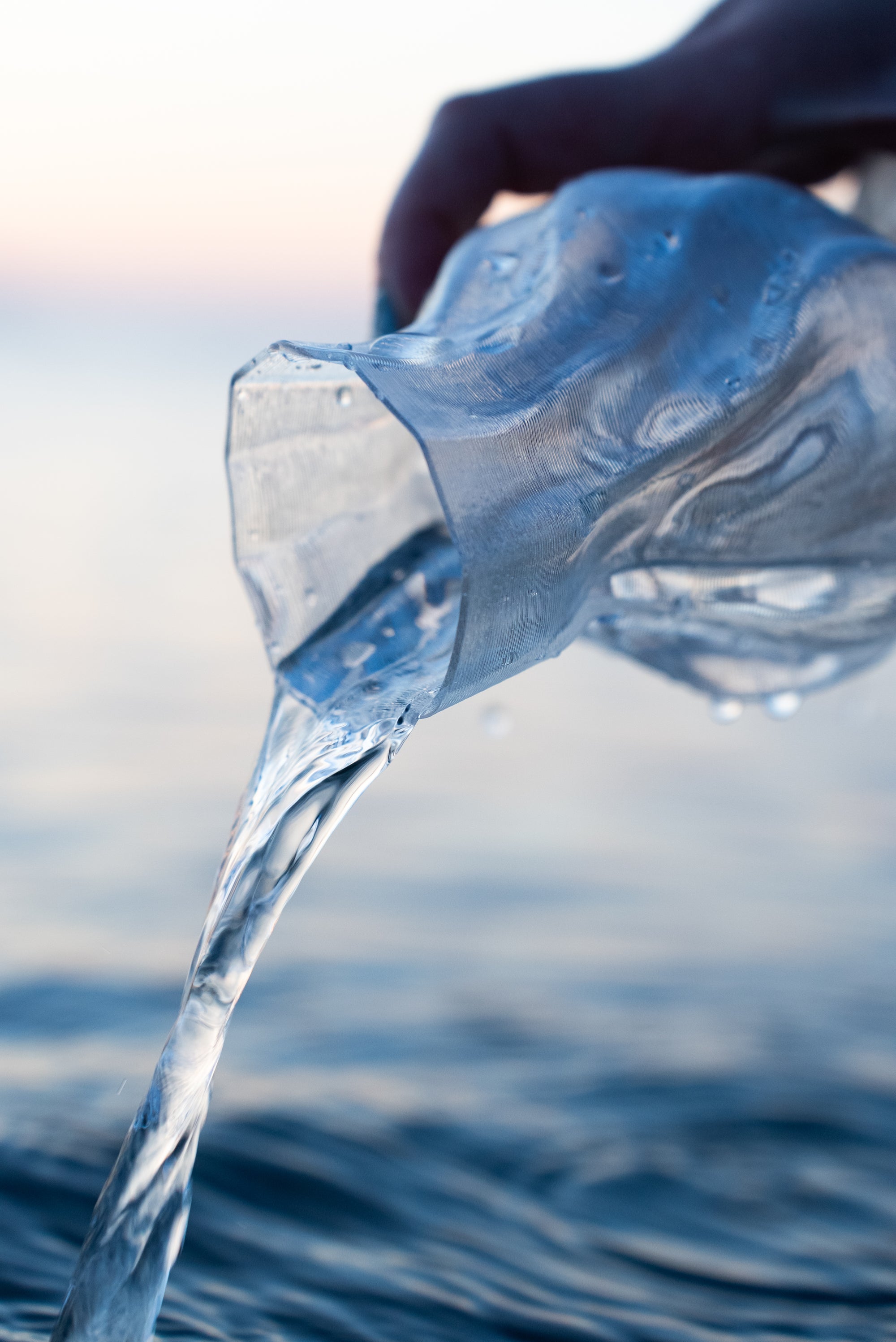 Main versant de l’eau à travers un vase Aria transparent au lever du soleil, au-dessus de la mer.