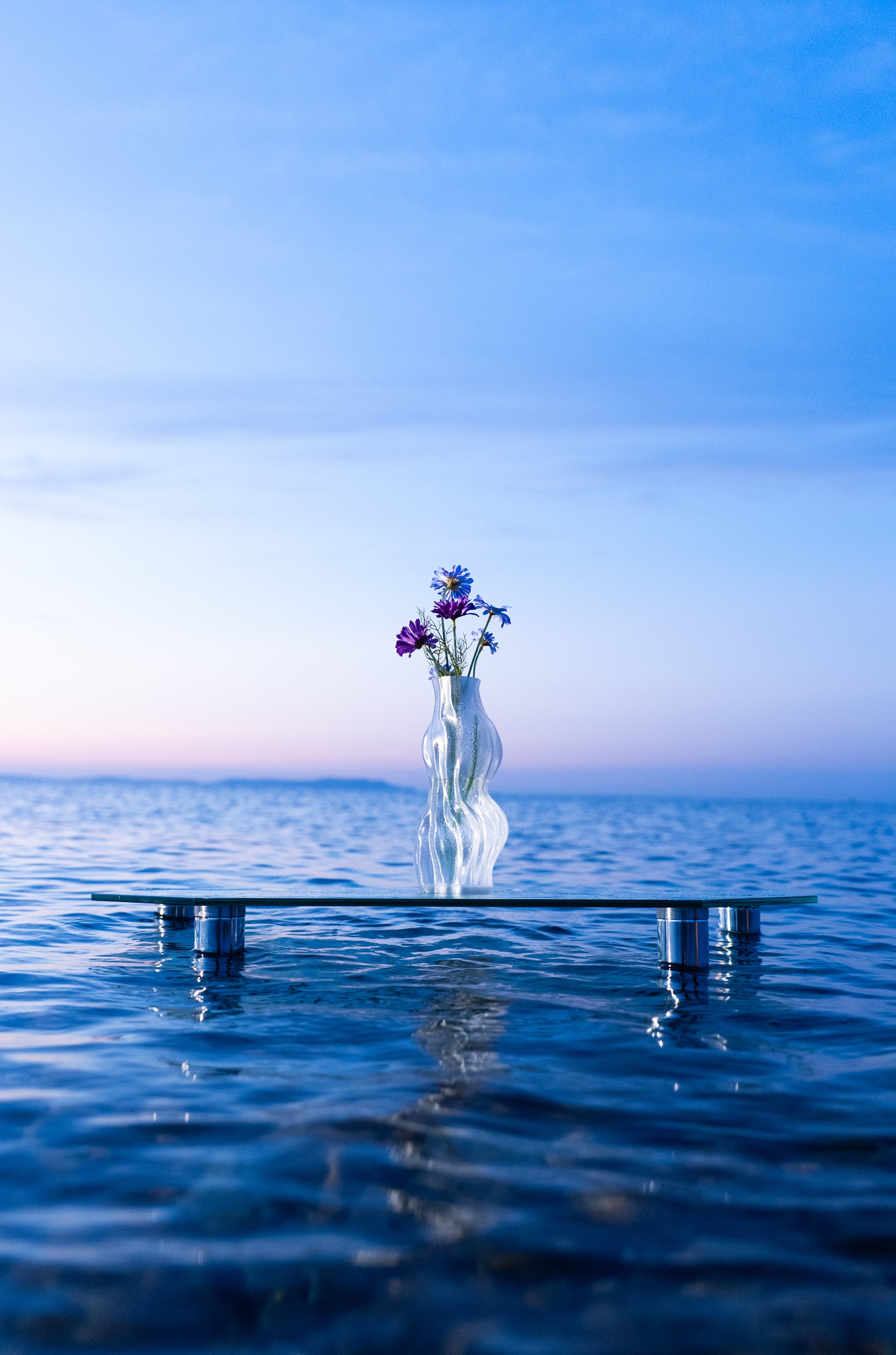Vase Aria transparent en PETG transparent, posé sur une table flottante au milieu de la mer au lever du soleil