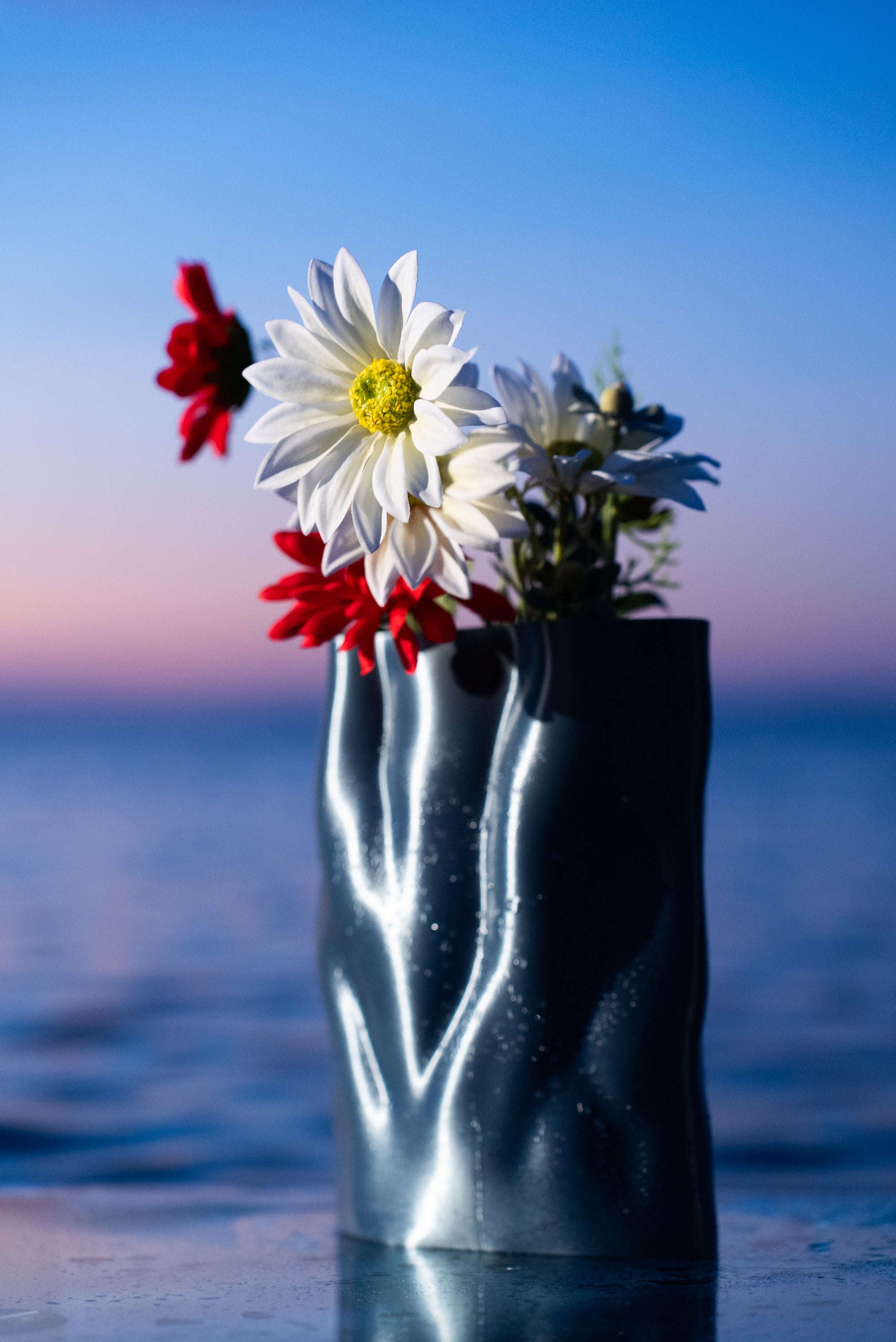 Vase Aura en PLA chromé de la collection Chrome Core, garni de fleurs, posé au bord de la mer au coucher du soleil