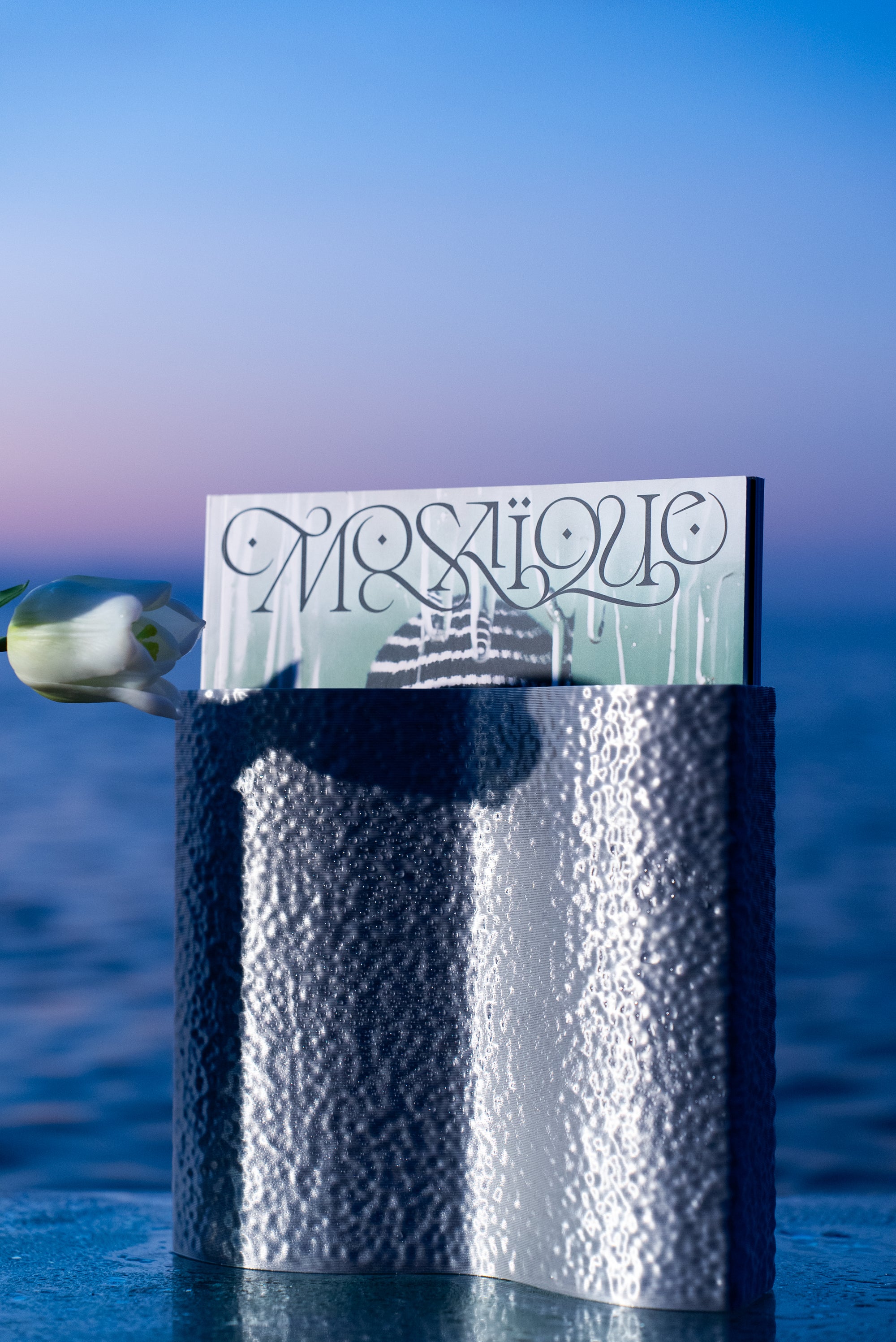 Vase Mira en PLA chromé à texture martelée, utilisé comme porte-magazine, au lever du soleil au bord de la mer
