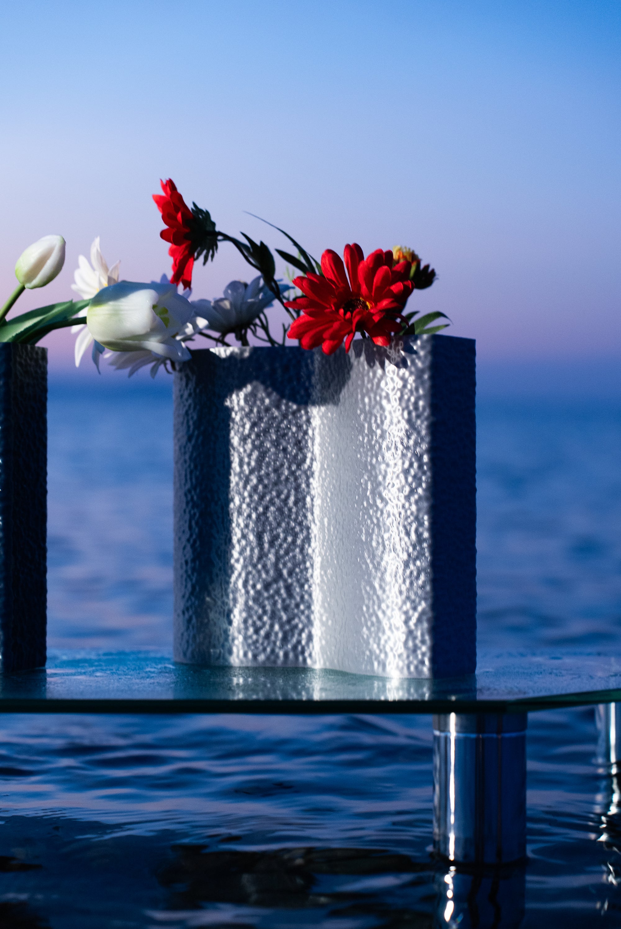 Vase Mira en coloris Chrome de S7DE Studio, garni de fleurs rouges et blanches, posé sur une table en verre au bord de la mer à l’aube.