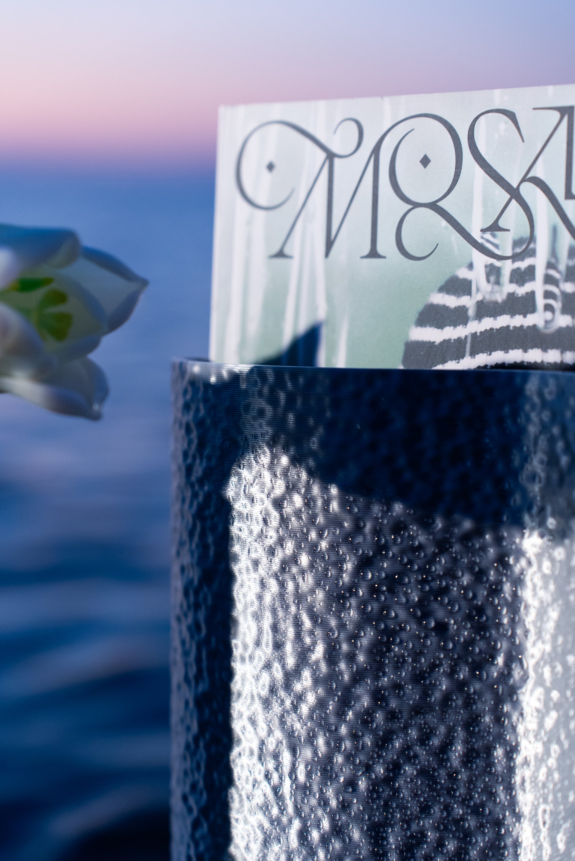 Vase Mira en coloris Chrome de S7DE Studio utilisé comme porte-magazine, contenant un exemplaire du magazine Mosaïque, dans une mise en scène au lever du soleil en bord de mer.