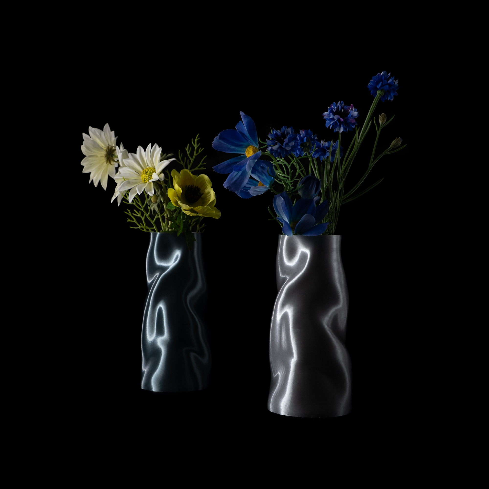 Deux vases Riflesso photographiés sur fond noir, remplis de fleurs fraîches. À gauche le modèle Aether avec des fleurs blanches et jaunes, à droite le modèle Chrome avec des fleurs bleues.