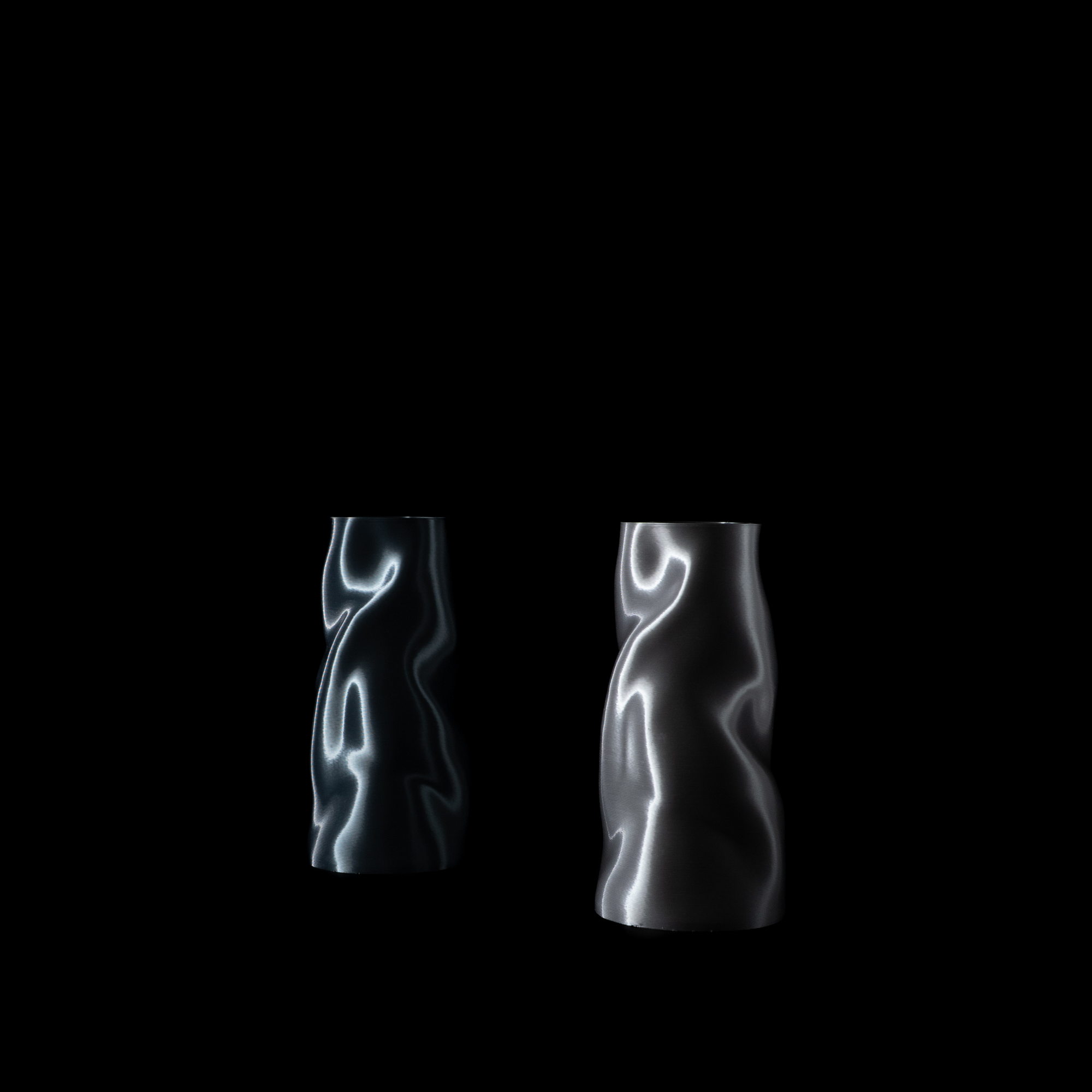 Deux vases Riflesso photographiés de face sur fond noir. À gauche le coloris Aether au ton plus froid, à droite le coloris Chrome plus lumineux.