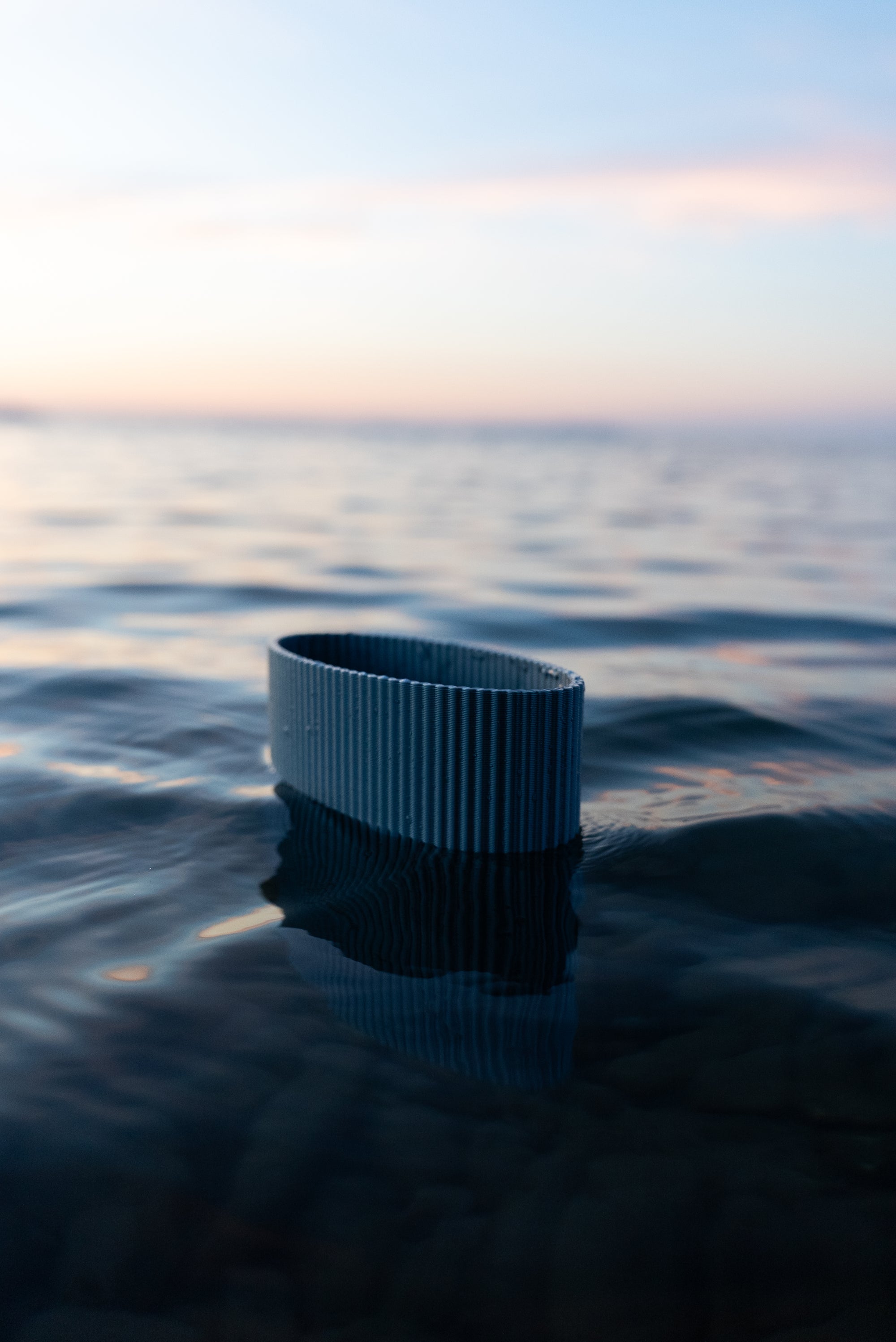 Vase Riga de S7DE Studio en coloris Aether flottant sur l’eau au coucher du soleil, avec un effet miroir subtil et des reflets ondulants.