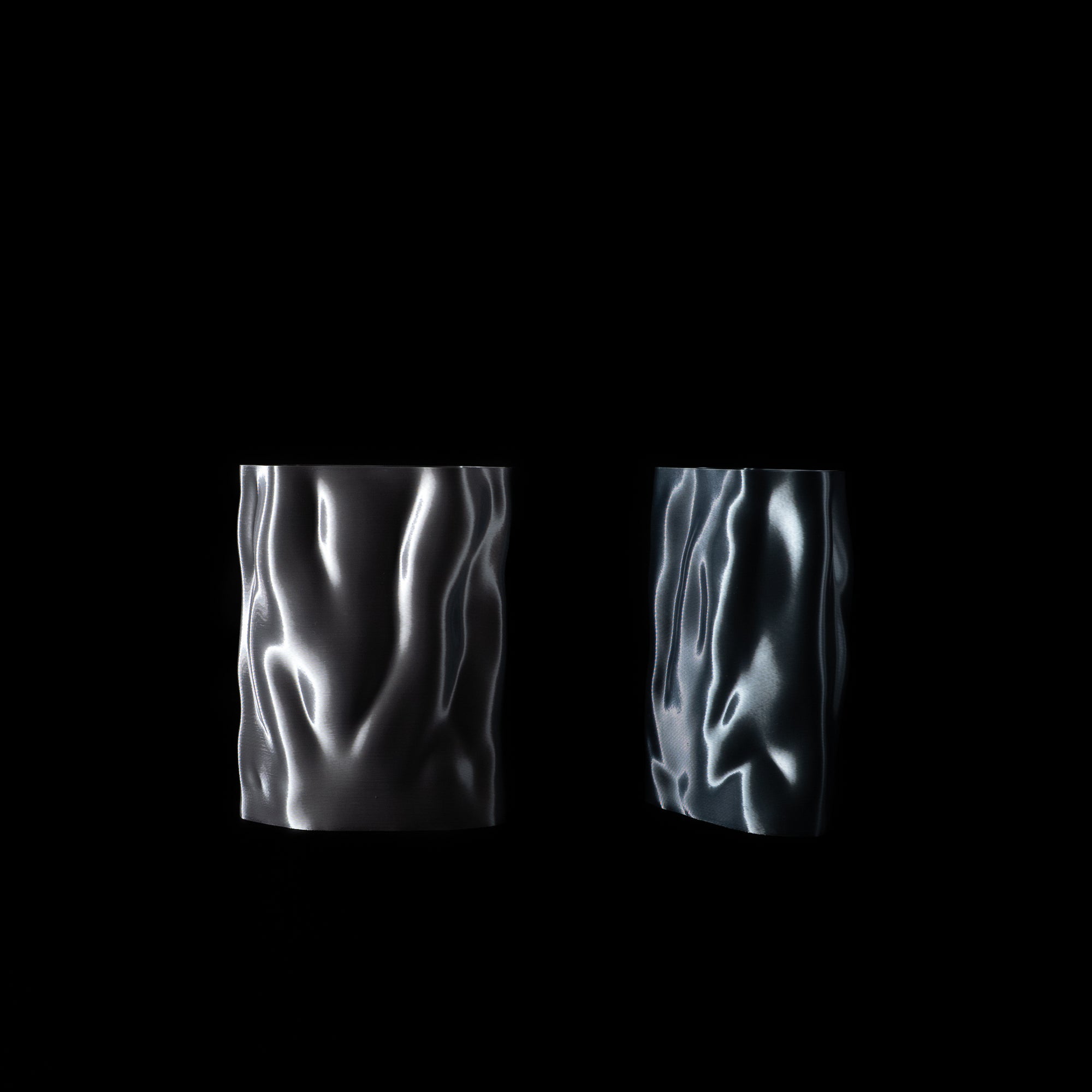 Deux vases Aura sur fond noir : à gauche en finition Chrome, à droite en finition Aether. Reflets métalliques et texture fluide mis en valeur par l’éclairage.