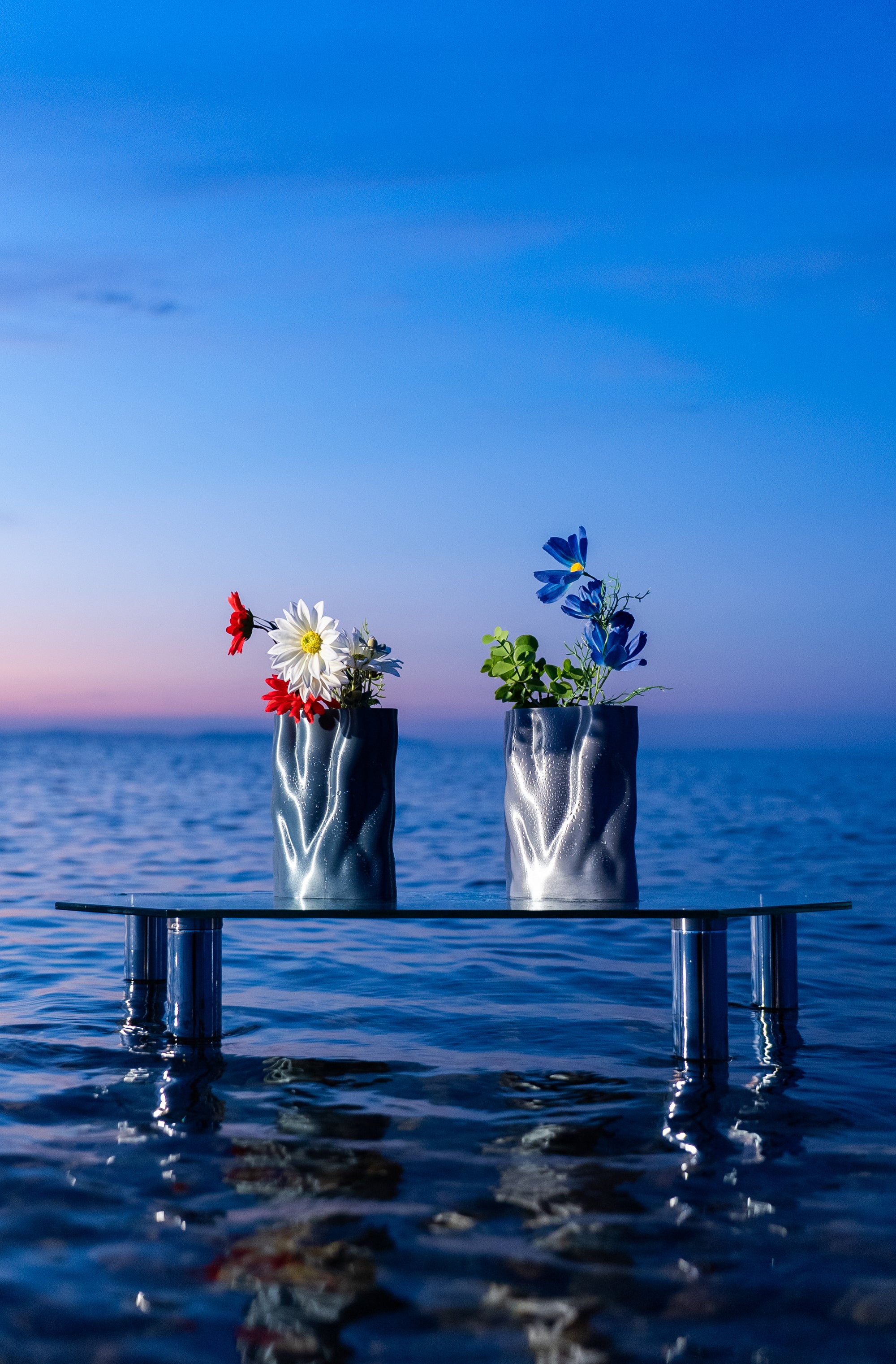 Duo de vases Aura de S7DE Studio flottant sur une table en verre posée sur la mer au lever du soleil, en coloris Aether (gauche) et Chrome (droite), ornés de bouquets de fleurs.