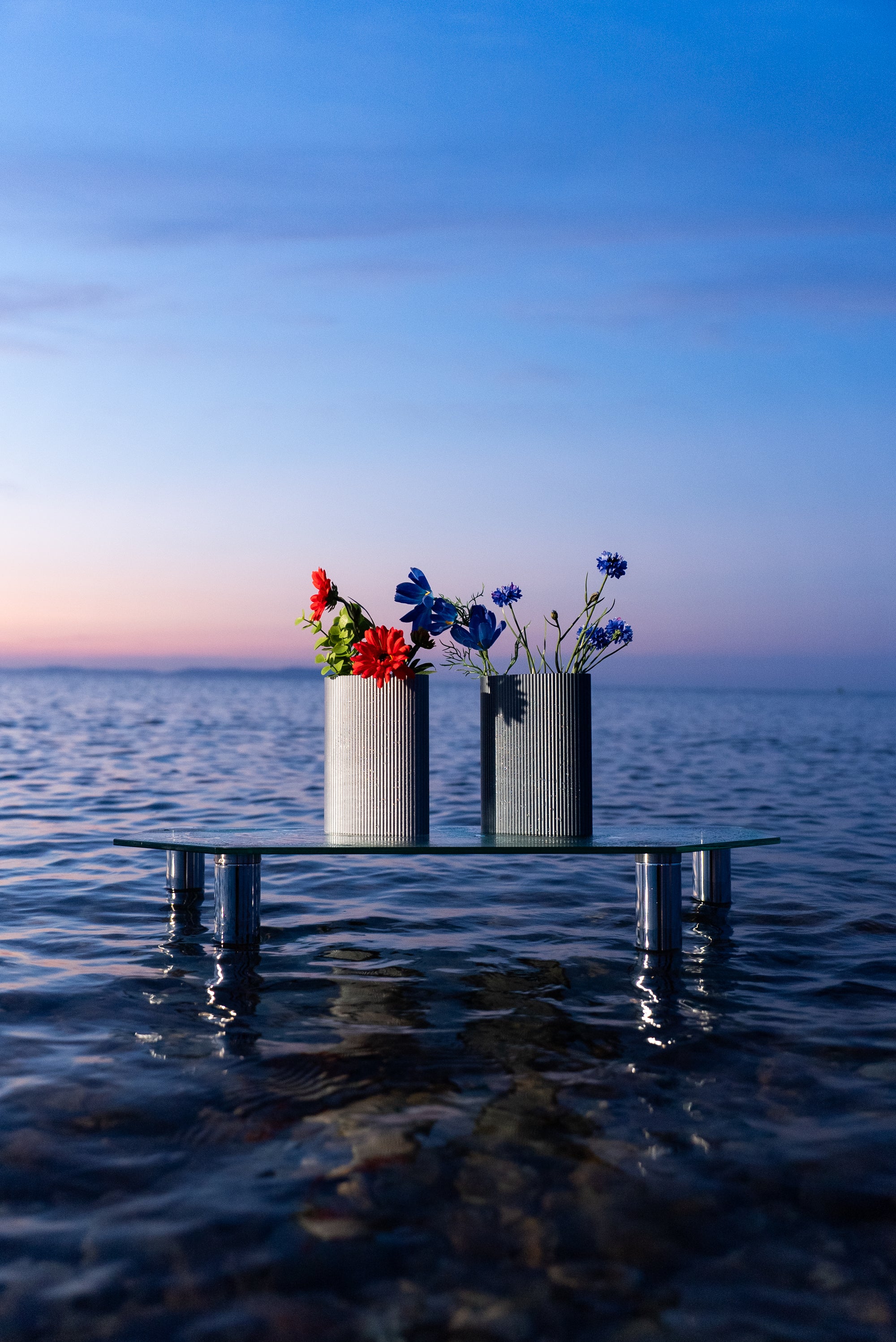 Deux vases Riga de S7DE Studio au lever du soleil, posés sur une table en verre dans la mer : coloris Chrome à gauche et Aether à droite, ornés de fleurs fraîches.