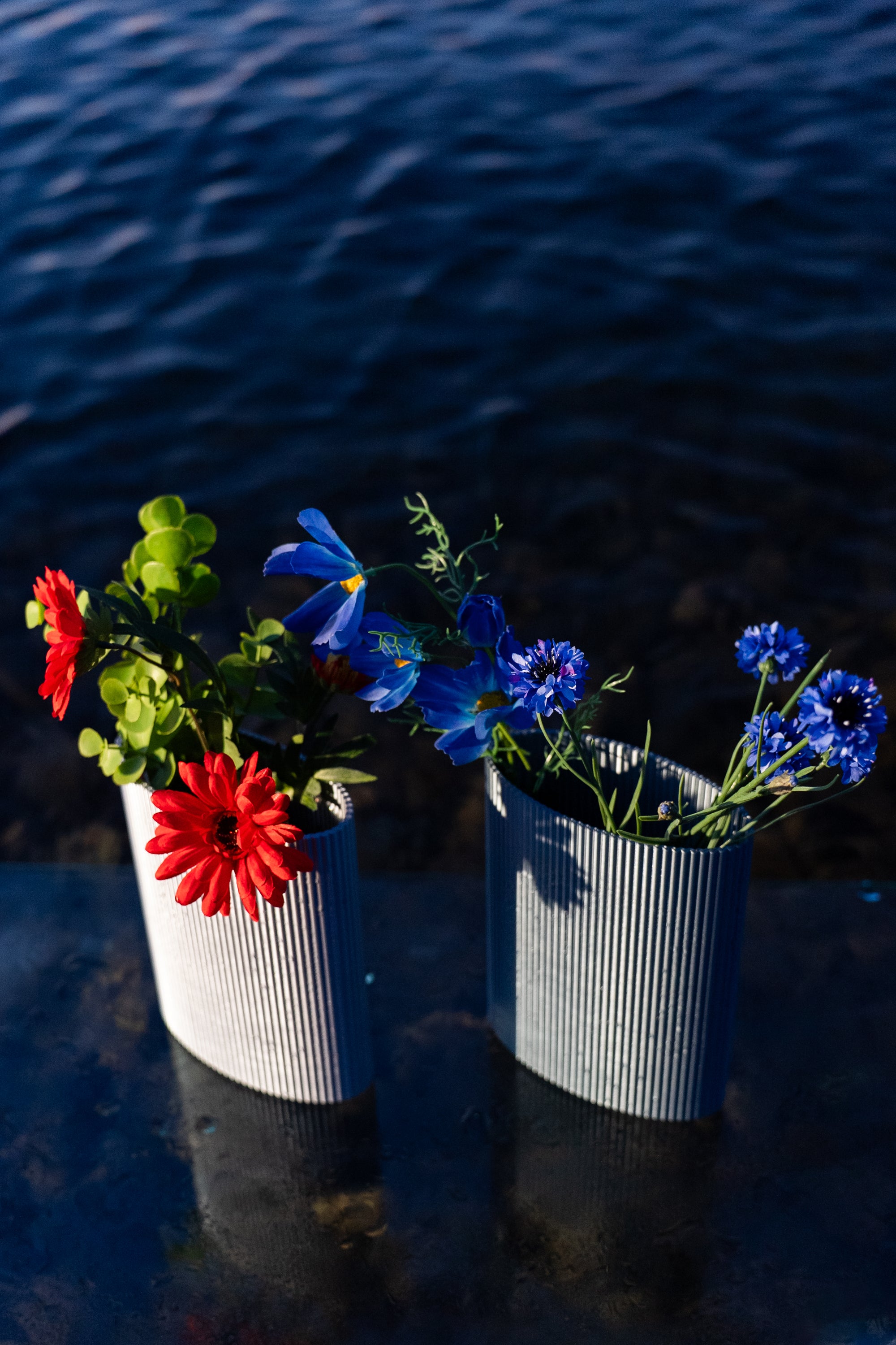 Duo de vases Riga en coloris Chrome et Aether de S7DE Studio, posés sur une table en verre au-dessus de l’eau, ornés de fleurs rouges et bleues.